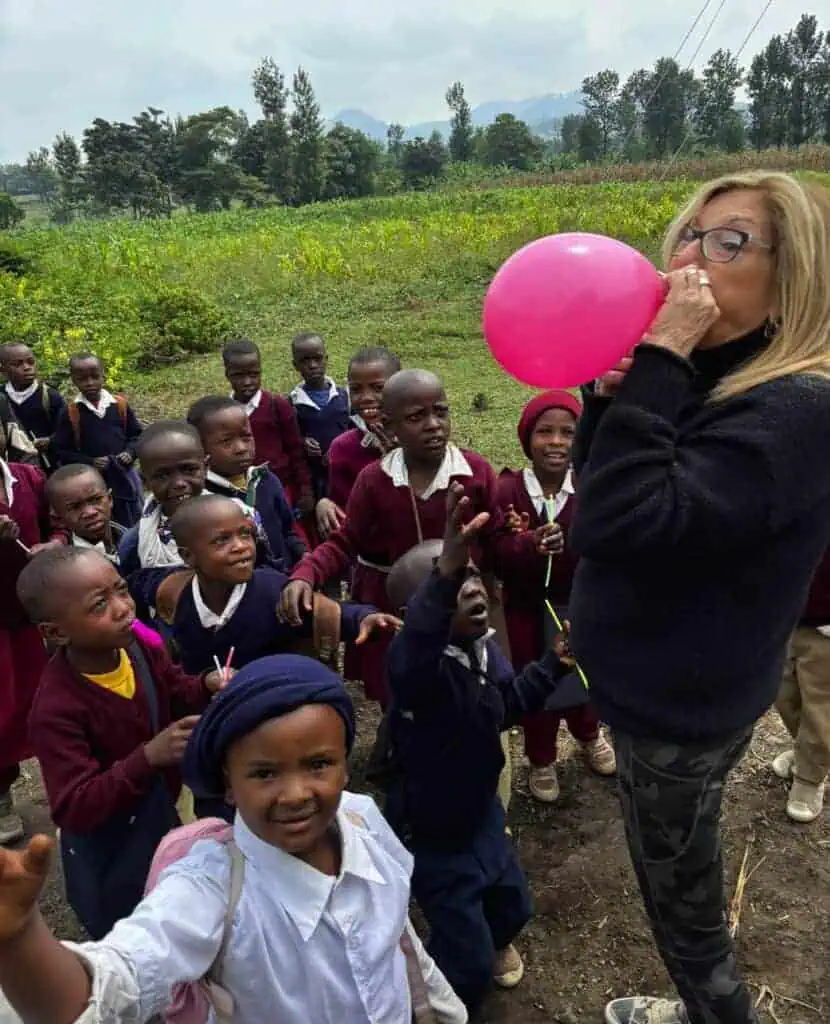 Pink balloon being blown by a woman to a group of excited schoolchildren outdoors.