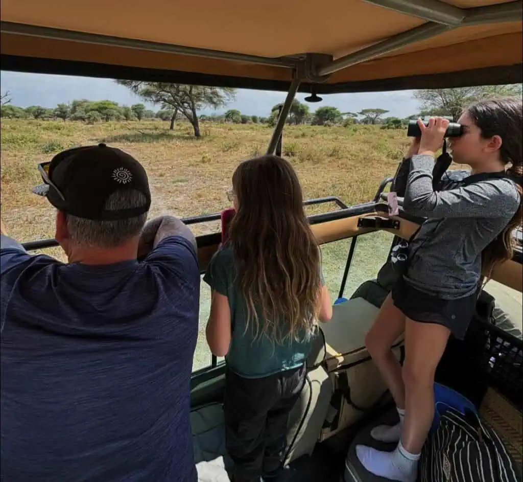 Wildlife safari adventure with children in open-sided vehicle, observing African savannah and wildlife, binoculars, family travel, guided tour, nature exploration, Africa safari experience, scenic landscape, and wildlife viewing.