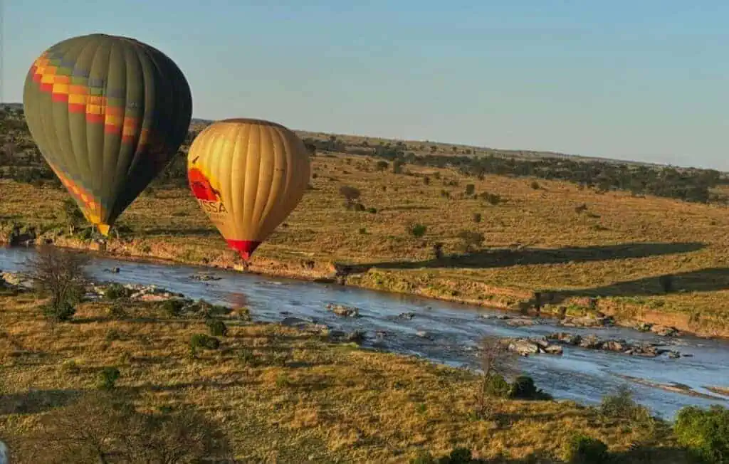 Hot air balloons floating over the African savannah during a safari adventure, with a river and open plains in the background, offering a breathtaking view of wildlife and landscapes.