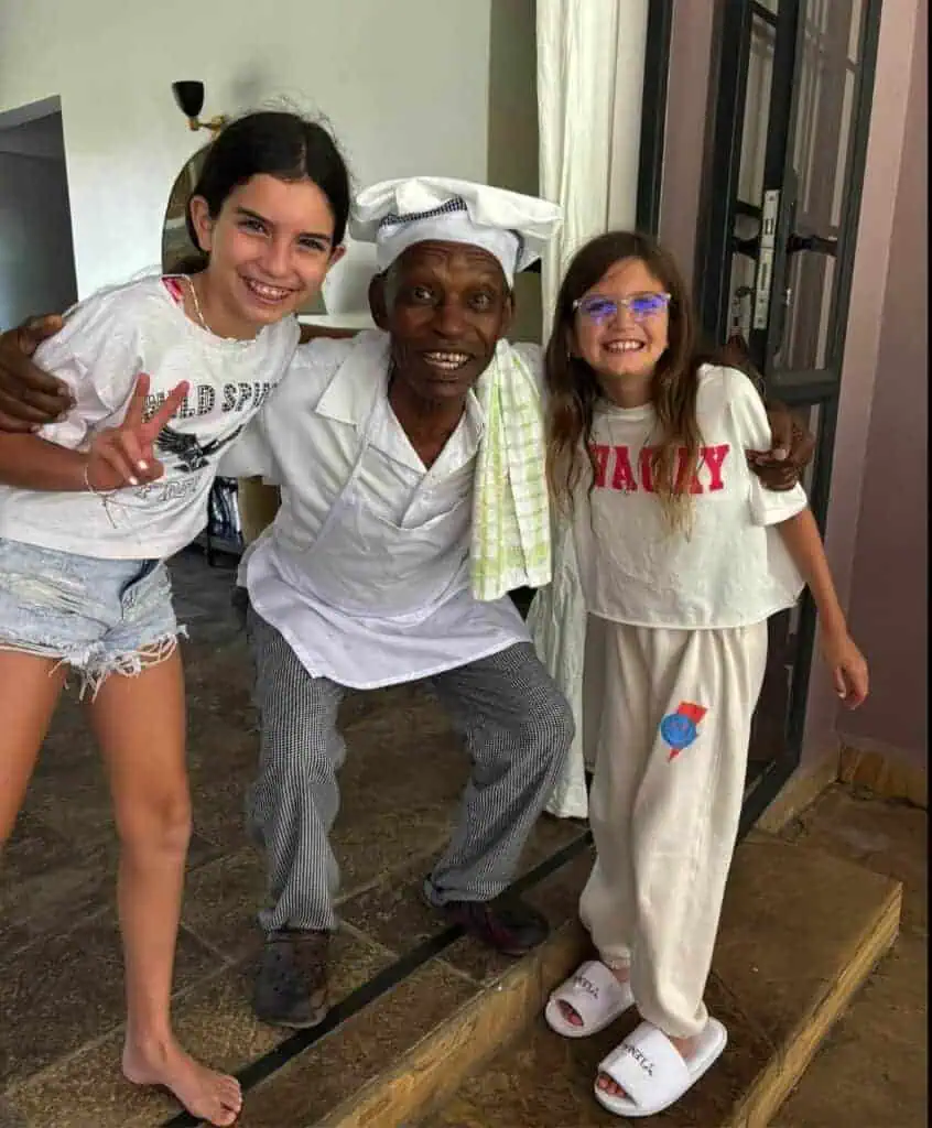 Joyful children and an elderly chef smile happily together in a cozy indoor setting, illustrating warm hospitality and cultural exchange in Africa.