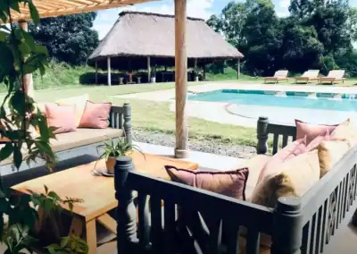 Comfortable outdoor seating area with cushions overlooking a swimming pool and traditional thatched hut in a scenic African setting.