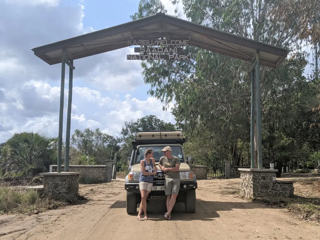 Me and my husband leaning on our safari vehicle beneath the entrance gate to Saadani National Park, Tanzania