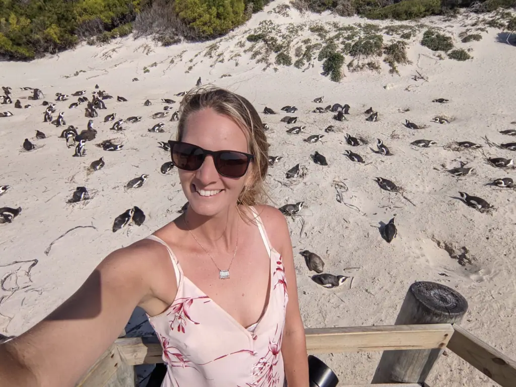 Me smiling in sunglasses on the sand at Boulders Beach, South Africa, with African penguins on the beach behind me